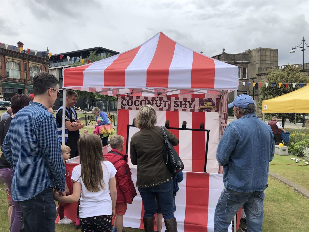 Players at the throwing line at a community event, with the striped coconut shy stall behind