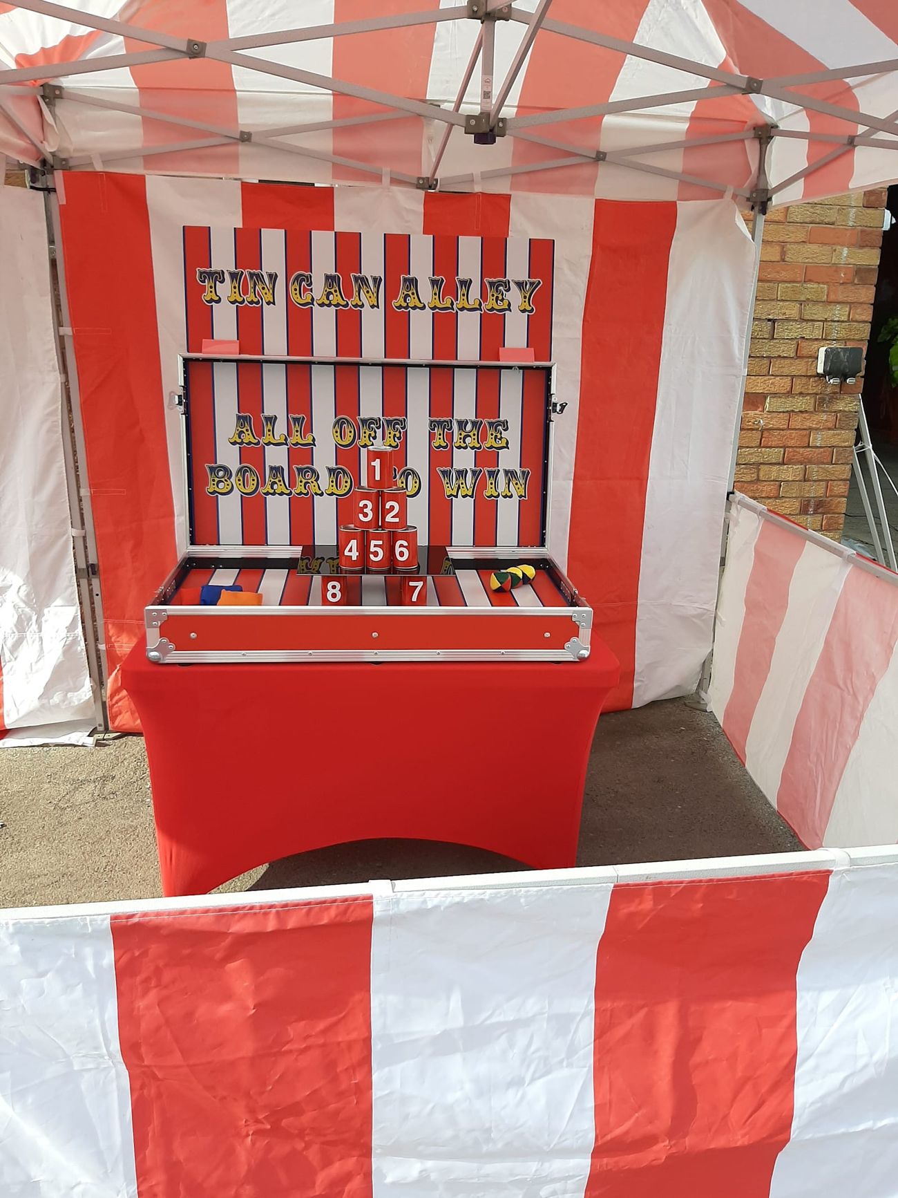 The Tin Can Alley flight case opened for play inside the striped stall, with the backboard and cans in place