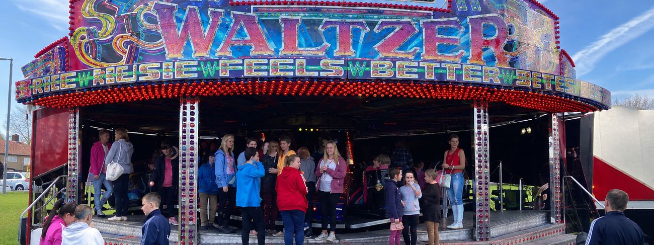 Harniess Waltzer set up on grass in daylight, with its full illuminated sign and a crowd gathered across the stepped loading deck